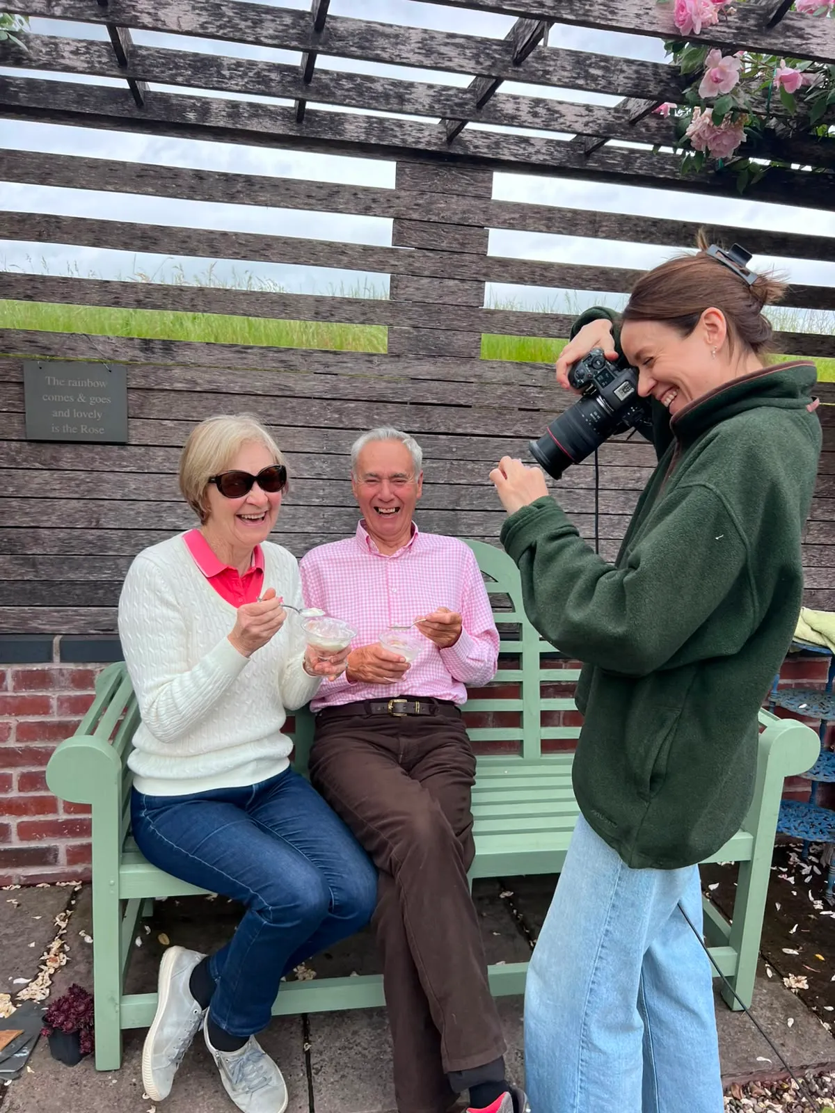 Behind the scenes: me photographing my parents laughing on a garden bench under the rose pergola, both eating ice cream from glass dishes.
