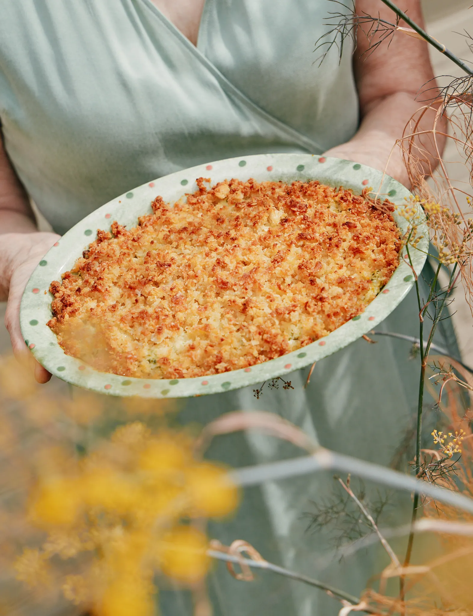 My mother holding out a spotted green dish of courgette crumble, golden breadcrumb topping just out of the oven, fennel flowers in the foreground.
