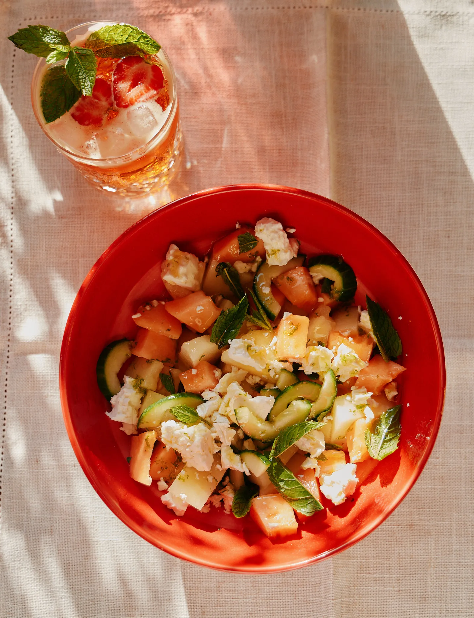 Watermelon, melon, cucumber, feta and mint salad in a red bowl beside an iced drink garnished with strawberry and mint.